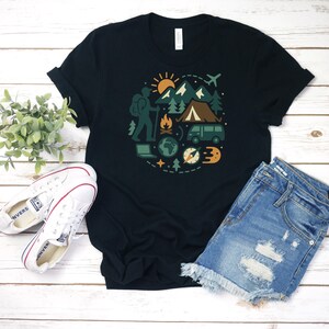 May include: Black t-shirt featuring a circular graphic of outdoor adventure elements. The design includes a hiker, mountains, a tent, a campfire, a van, and a compass. The shirt is paired with white sneakers and denim shorts.