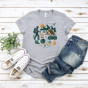 May include: A heather gray t-shirt featuring a circular graphic of outdoor adventure elements. The design includes a hiker, mountains, a tent, a van, and a compass. The shirt is paired with blue jeans and white sneakers.