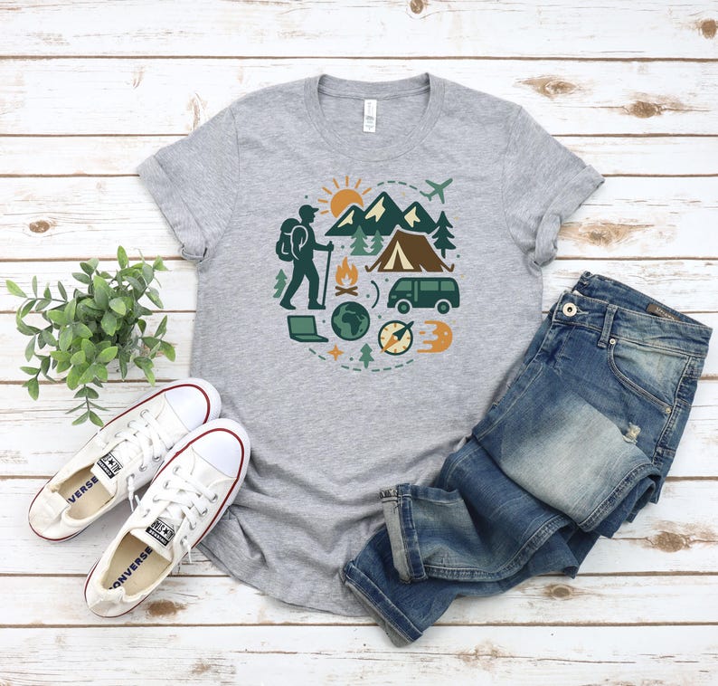 May include: A heather gray t-shirt featuring a circular graphic of outdoor adventure elements. The design includes a hiker, mountains, a tent, a van, and a compass. The shirt is paired with blue jeans and white sneakers.