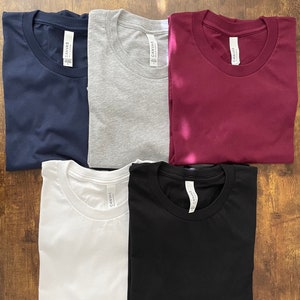 May include: A collection of six folded t-shirts in various colors: navy blue, gray, burgundy, white, and black. Each shirt has a crew neck and a tag that reads "CANVAS". The shirts are arranged on a wooden surface.