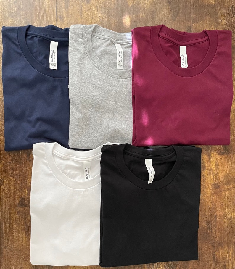 May include: A collection of six folded t-shirts in various colors: navy blue, gray, burgundy, white, and black. Each shirt has a crew neck and a tag that reads "CANVAS". The shirts are arranged on a wooden surface.
