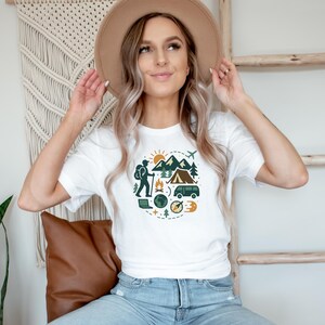 May include: White t-shirt featuring a circular graphic with camping and travel elements. The design includes mountains, a tent, a hiker, a compass, and a van. The person is wearing a tan hat and blue jeans.
