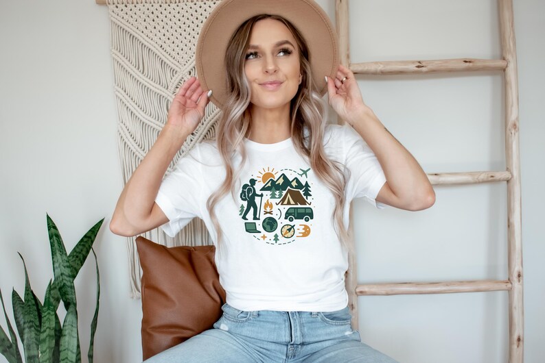 May include: White t-shirt featuring a circular graphic with camping and travel elements. The design includes mountains, a tent, a hiker, a compass, and a van. The person is wearing a tan hat and blue jeans.