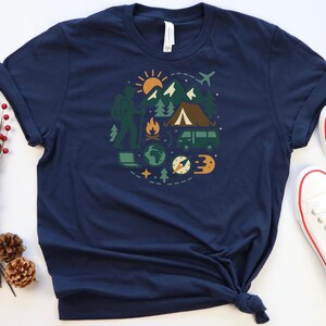 May include: Navy blue t-shirt featuring a circular graphic of outdoor adventure elements. The design includes a hiker, mountains, a tent, a van, a compass, and a sun. The shirt is made of a soft material.