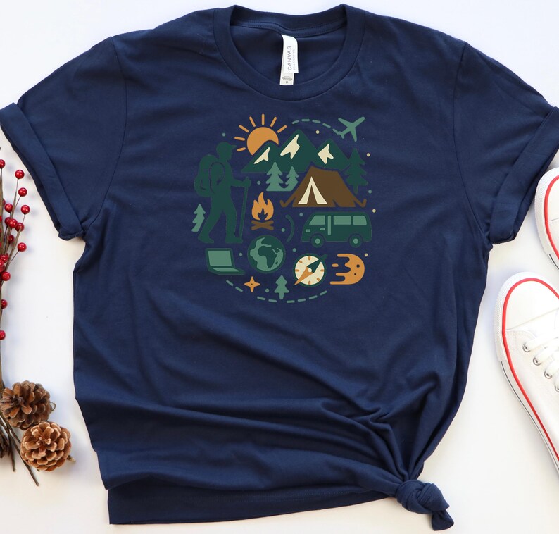 May include: Navy blue t-shirt featuring a circular graphic of outdoor adventure elements. The design includes a hiker, mountains, a tent, a van, a compass, and a sun. The shirt is made of a soft material.