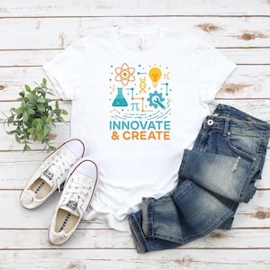 May include: White t-shirt with a colorful graphic design featuring an atom, lightbulb, and gear, with the words "Innovate & Create." The shirt is paired with blue jeans and white sneakers with red accents. The items are arranged on a light wood surface.