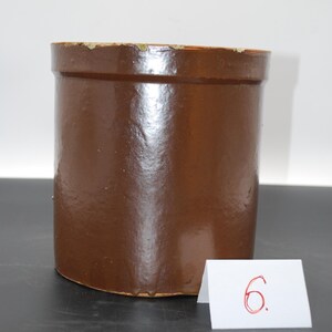 Puede incluir: Un tarro de cerámica marrón con forma cilíndrica. El tarro tiene un borde ligeramente ensanchado y un acabado liso y brillante. Una pequeña tarjeta blanca con el número "6" escrito en rojo está delante del tarro.