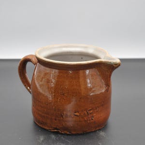 Op de afbeelding: Een bruine keramische kan met een wit geglazuurd interieur en een handvat. De kan heeft een rond lichaam en een licht uitlopende lip.