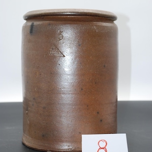 Puede incluir: Un tarro de gres marrón con tapa. El recipiente cilíndrico tiene un esmalte marrón oscuro y una marca de capacidad de "5" y un logotipo triangular. Una pequeña tarjeta blanca con el número "8" está frente al tarro.