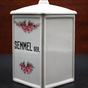 May include: A white ceramic storage canister with a square base and a matching lid. The front features the black text "SEMMEL GER." centered between two floral decals of pink roses with green leaves. The canister has a smooth, glossy finish.