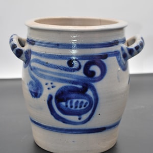 Könnte beinhalten: Eine weiße Keramik-Dose mit zwei Henkeln und blauen bemalten Designs. Die Dose hat eine breite Öffnung und einen runden Körper.