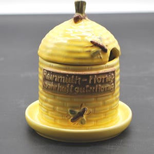 Könnte beinhalten: Gelber Keramik-Honigtopf in Bienenkorbform mit Deckel. Der Topf hat ein braunes rechteckiges Etikett mit deutschem Text. Eine kleine Untertasse befindet sich unter dem Topf. Dekorative Bienen sind auf den Topf gemalt. Der Topf ist ca. 10 cm hoch.