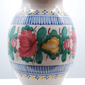 May include: A white ceramic vase with a wide body and a narrow neck. The vase is decorated with colorful floral patterns in red, pink, yellow, and green. Blue and yellow bands encircle the vase.