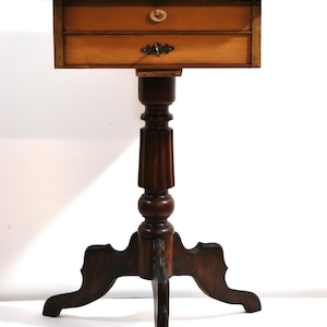 Könnte beinhalten: Ein Holz-Beistelltisch mit zwei Schubladen und einem gedrehten Säulenfuß. Der Tisch hat eine helle Holzplatte und einen dunklen Holzfuß.