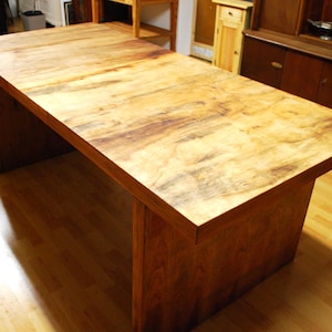 Könnte beinhalten: Ein großer, rechteckiger Holztisch mit natürlicher Maserung. Der Tisch hat ein solides, blockartiges Gestell und eine dicke Tischplatte. Die Holztöne reichen von hellbraun bis dunkelbraun. Der Tisch ist etwa 183 cm lang.