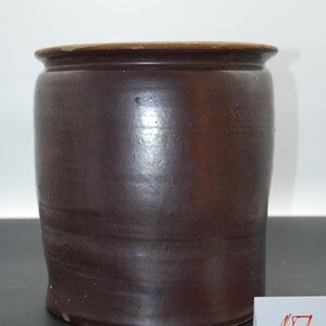 Op de afbeelding: Een donkerbruine steengoed kruik met een licht uitlopende rand. De cilindrische container heeft een gladde, glanzende afwerking en een lichtere bruine rand. Het nummer "17" staat op een kleine witte kaart in de rechter benedenhoek.