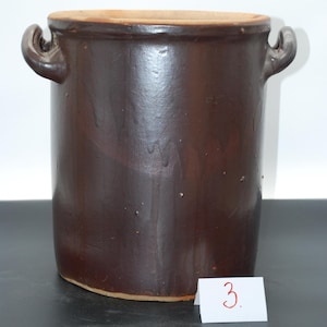 May include: A dark brown ceramic crock with two curved handles. The cylindrical container has a wide opening and a lighter brown rim. A small white card with the number "3" is visible.