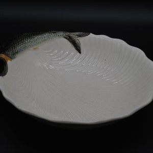 Könnte beinhalten: Weiße Keramikschüssel mit Fischdesign. Die Schüssel hat einen gewellten Rand und eine strukturierte Oberfläche. Der Fisch ist in Grautönen und Schwarz bemalt.