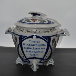 Può includere: Un vaso di ceramica bianca con un motivo floreale blu e verde. Il vaso ha un coperchio con un pomo in cima. Il vaso ha una citazione in tedesco sul davanti: "Tropfen des Geistes Giesset hinein, Leben dem Leben gibt er allein."