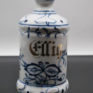 Könnte beinhalten: Eine weiße Keramik-Dose mit blauen und goldenen Blumendesigns. Die Dose hat eine goldene Aufschrift mit der Aufschrift "Essig".