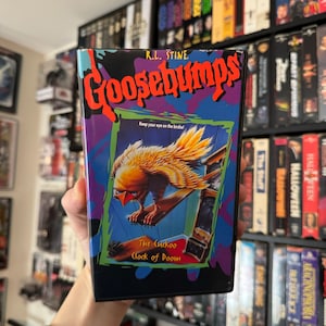 May include: A Goosebumps VHS tape featuring a colorful illustration of a bird perched on a clock. The title "The Cuckoo Clock of Doom" is visible, along with the author's name, R.L. Stine. The tape is set against a backdrop of other VHS tapes.