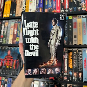 May include: VHS tape case for the horror movie "Late Night with the Devil". The cover features a man in a suit standing in flames with a dark background. The title is in white bold letters.