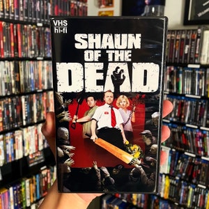 May include: A VHS tape case for the movie "Shaun of the Dead". The case is black with a red background and features the movie title in white text. The image shows the main characters from the movie, including Shaun, Ed, and Liz, standing in front of a crowd of zombies.