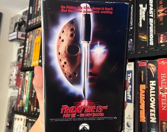 May include: A VHS tape for the film Friday the 13th Part VII: The New Blood. The cover features a close-up of Jason's hockey mask and a knife going through a woman's eye. The text on the cover reads "On Friday the 13th, Jason is back. But this time someone's waiting."