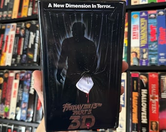 Può includere: Una videocassetta VHS del film "Venerdì 13, Parte 3: 3D". La copertina presenta un'immagine in bianco e nero di Jason Voorhees in piedi dietro una tenda da doccia, con un coltello in mano. Il testo "A New Dimension In Terror..." è in cima alla copertina.