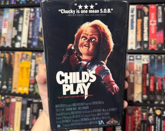 May include: A VHS tape for the horror film "Child's Play" featuring a red-haired doll with a knife. The movie poster features the quote "Chucky is one mean S.O.B." -ROGER EBERT, New York Daily News.
