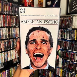 以下が含まれることがあります： 映画「アメリカン・サイコ」のVHSテープケース。血まみれの顔をしたパトリック・ベイトマンのアップが写っている。ケースには「VHS hi-fi」と「Bret Easton Ellisの物議を醸すベストセラーから」という文字が印刷されている。