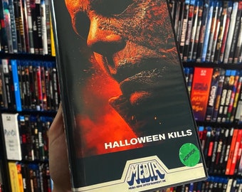 Może przedstawiać: Kaseta VHS z filmem grozy "Halloween Kills" z bliskim ujęciem twarzy zamaskowanej postaci. Tytuł jest w białych literach blokowych na czarnym tle, z zieloną naklejką "HORROR".
