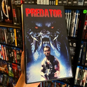 May include: A VHS tape case with a blue and black image of a Predator creature and a man holding a gun. The text "PREDATOR" is in red letters on the front of the case.