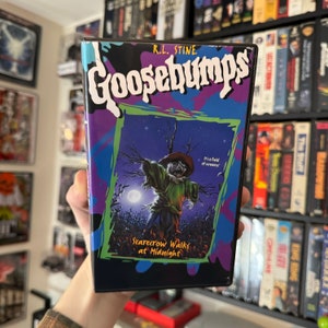 May include: A Goosebumps VHS tape titled "Scarecrow Walks at Midnight." The cover features a scarecrow illustration against a night sky background. The title is in large, white, stylized letters with a purple and blue border.