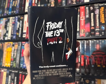 May include: A VHS tape for the film "Friday the 13th Part 2". The cover features a black silhouette of a person holding an axe with blood dripping from the axe. The text "The body count continues..." is printed below the silhouette.