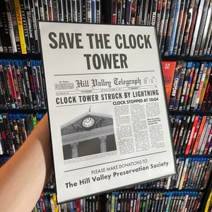 May include: A black and white newspaper clipping with the headline "Clock Tower Struck By Lightning" and the text "Save The Clock Tower". The newspaper is from the "Hill Valley Telegraph" and is dated November 14, 1955.