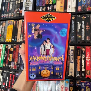 Może przedstawiać: Kaseta VHS z filmem Halloweentown z serii Klasyki Disneya. Okładka jest czerwona z kreskówkowym obrazkiem miasta z dynią na szczycie budynku. Tekst "Halloweentown" jest w kolorze żółtym, pomarańczowym i czerwonym. Tekst "Walt Disney Classics" jest w kolorze białym.