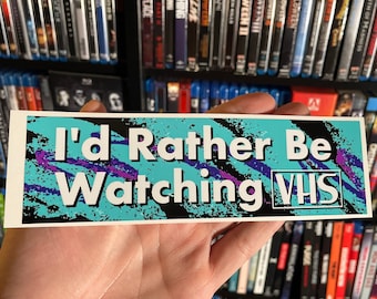 May include: A sticker with a blue, black, and purple 90s-style graphic design. The sticker says "I'd Rather Be Watching VHS" in white text.