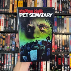 May include: A VHS tape of Stephen King's Pet Sematary. The cover features a green and black design with a close-up of a man's face and a cat in a graveyard. The title is in white and red lettering.