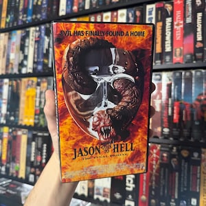 Op de afbeelding: Een VHS-bandhoes voor de horrorfilm "Jason Goes to Hell: The Final Friday". De hoes toont een close-up van Jason's hockeymasker met een worm-achtig wezen erin. De achtergrond is een vurig oranje en rood.