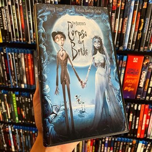 May include: A DVD cover for the animated film "Corpse Bride" by Tim Burton. The cover features the main characters, a pale-skinned groom and a skeletal bride, standing in a dark, gothic setting. The title "Corpse Bride" is written in a stylized font.
