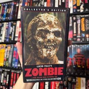 Könnte beinhalten: Eine VHS-Kassette für den Film "Zombie" von Lucio Fulci. Das Cover zeigt eine Nahaufnahme eines verfallenden Zombiegesichts in Braun- und Beigetönen. Der Titel "ZOMBIE" ist rot, mit dem Text "COLLECTOR'S EDITION" oben.