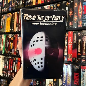 Puede incluir: Una cinta VHS de "Viernes 13 Parte V: Un Nuevo Comienzo". La portada presenta una máscara de hockey blanca con un círculo rosa sobre un fondo oscuro. El título está en blanco, con el título de la película arriba.