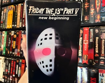 May include: A VHS tape for "Friday the 13th Part V: A New Beginning." The cover shows a white hockey mask with a pink circle against a dark backdrop. The title is in white, with the film's title at the top.