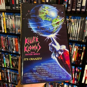 May include: A VHS tape cover for the horror movie "Killer Klowns from Outer Space". The cover features a clown's hand holding a glowing orb that is a cracked version of the Earth. The text on the cover reads "Killer Klowns from Outer Space" and "It's Craazzy!"