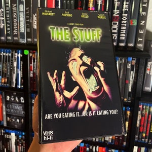 Puede incluir: Carátula de cinta VHS para la película de terror "The Stuff" con un hombre con una expresión de horror y una sustancia verde que le gotea de la boca. El eslogan dice "Are you eating it...or is it eating you?"