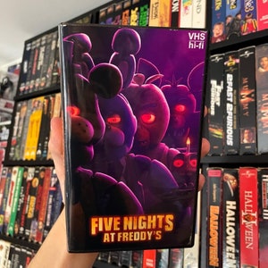 May include: A VHS tape for "Five Nights at Freddy's" featuring a purple and black cover with character illustrations and the title in gold. The tape is labeled "VHS hi-fi".