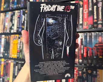 May include: A black VHS tape case featuring a dark image of a figure holding a knife. The title "FRIDAY THE 13TH" is printed in white on the case. The back of the case lists the cast and crew of the film.