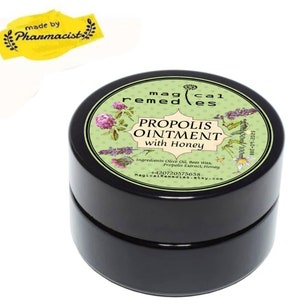 Op de afbeelding: Een zwarte, ronde pot "PROPOLIS OINTMENT with Honey" met een groen en wit etiket. Het etiket bevat bloemenillustraties en de tekst "magical remedies". Een gele sticker leest "made by Pharmacist."