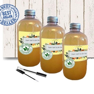 Puede incluir: Tres botellas transparentes llenas de un líquido dorado, etiquetadas como "Organic Liquid Lip gloss base". Cada botella tiene una etiqueta amarilla con diseños de naranja y flores y un sello de "100% Natural Ingredients". Dos aplicadores están frente a las botellas. Un sello de "Best Seller" está en el fondo.